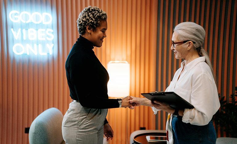 Two people shaking hands at the end of a meeting with a sign reading "Good vibes only" in the background.