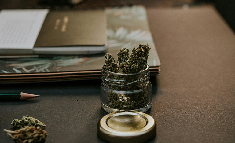 A bunch of marijuana flower in a glass jar sitting on a desk