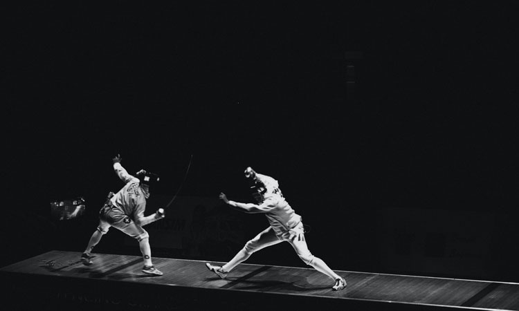 Two people fencing in white uniforms in a dark room