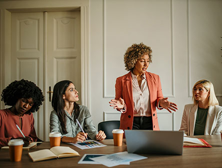Four professional women working at a large table while one speaks to the group.