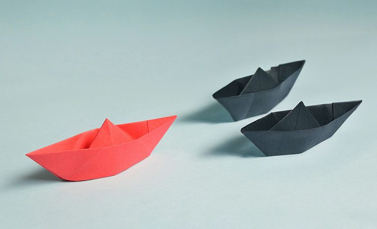 A red paper boat in front of two black paper boats