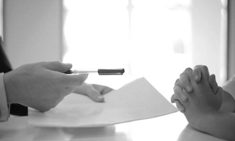 Person's hands holding a pen and paper in front of another set of hands clasped together