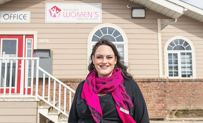 Danielle Moore, Family Law Advocate, stands in front of Fort St. John Women’s Resource Society. She wears a bright fuchsia scarf over a black top and smiles towards the camera.