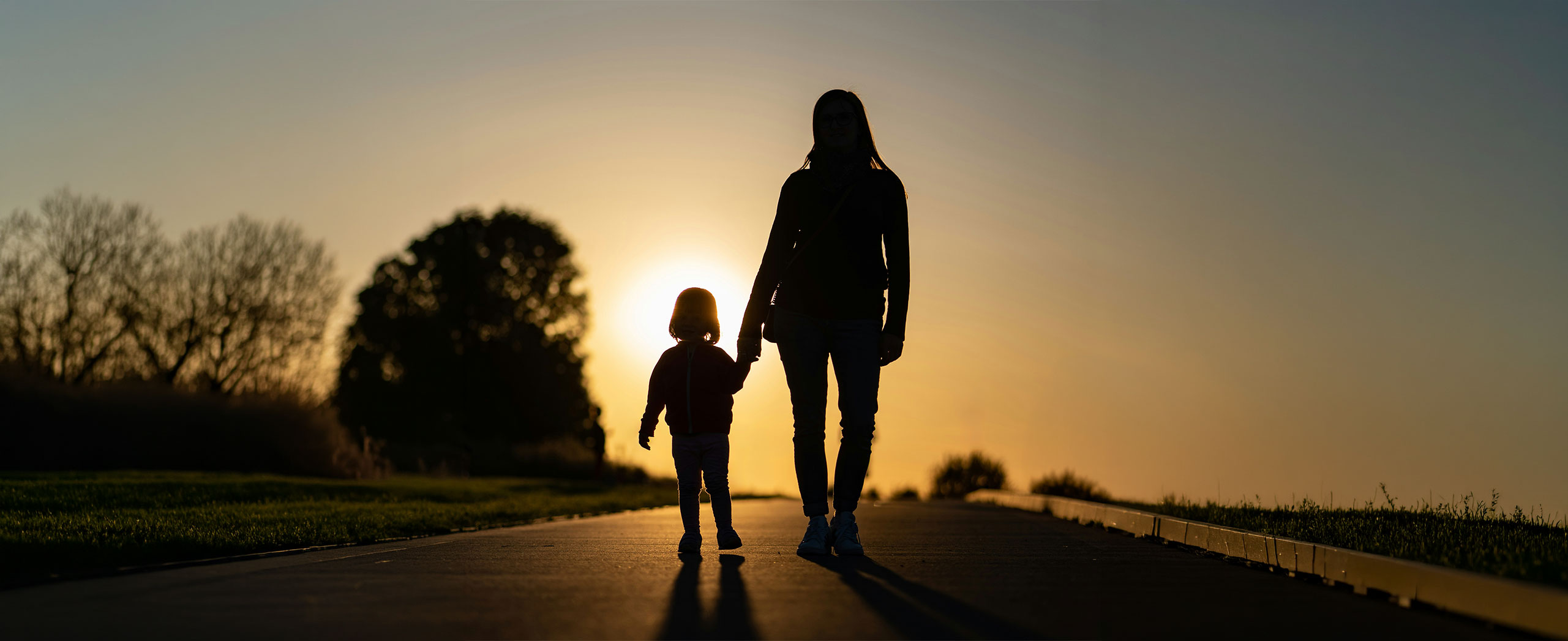 A mother walking and holding her child's hand as they walk towards a setting sun.
