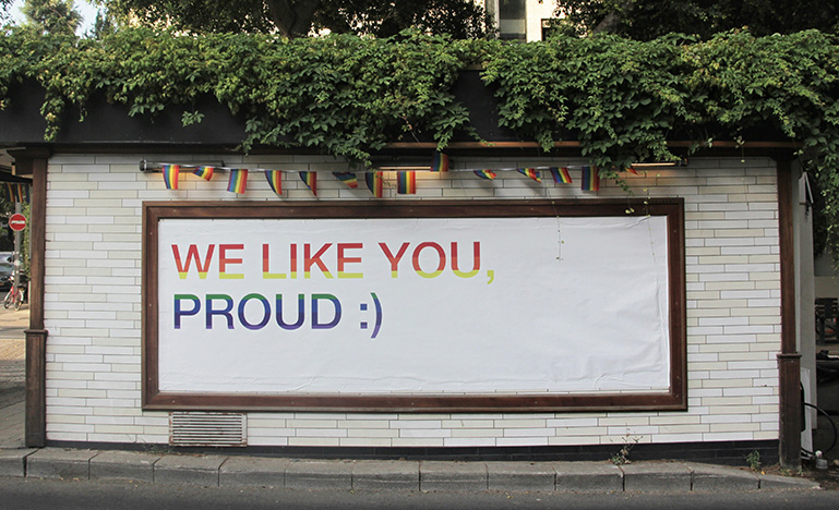 A printed sign in rainbow colours that says "We like you proud :)"