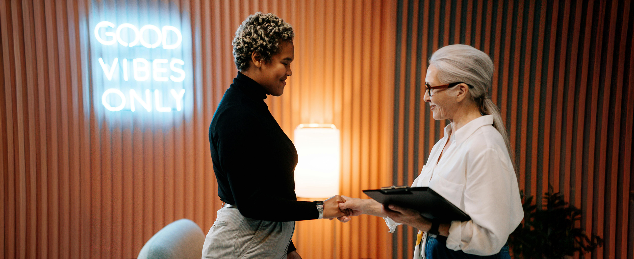 Two people shaking hands at the end of a meeting with a sign reading "Good vibes only" in the background.