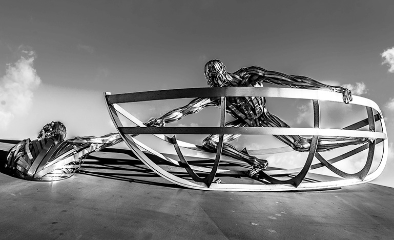 Silver mental sculpture of a person in a boat frame holding the arm of another statue of person waist deep in the ground