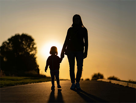 A mother walking and holding her child's hand as they walk towards a setting sun.