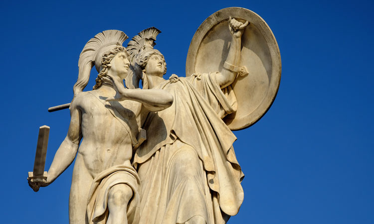 Two beige statues of Greek soldiers holding a sword and shield
