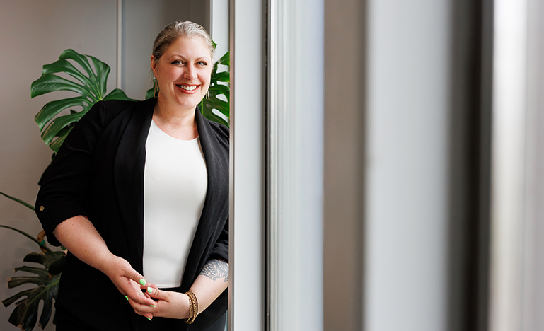 Maggie House leans against a wall at her workplace and smiles towards the camera. She is wearing a white top and black jacket.